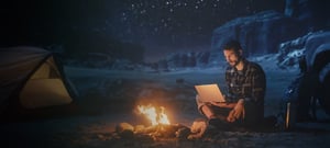 Man working on a laptop at campsite in front of a fire