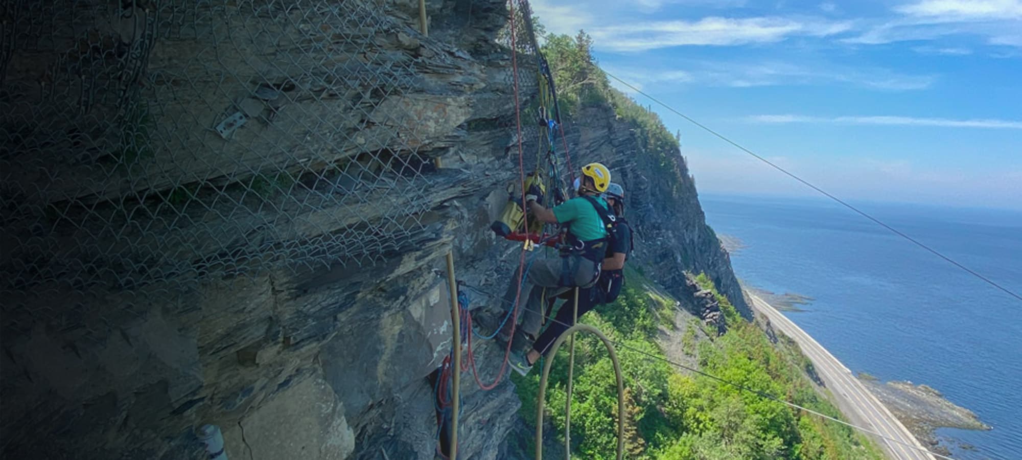 Two technicians in climbing harnesses installing equipment on the side of a rock face